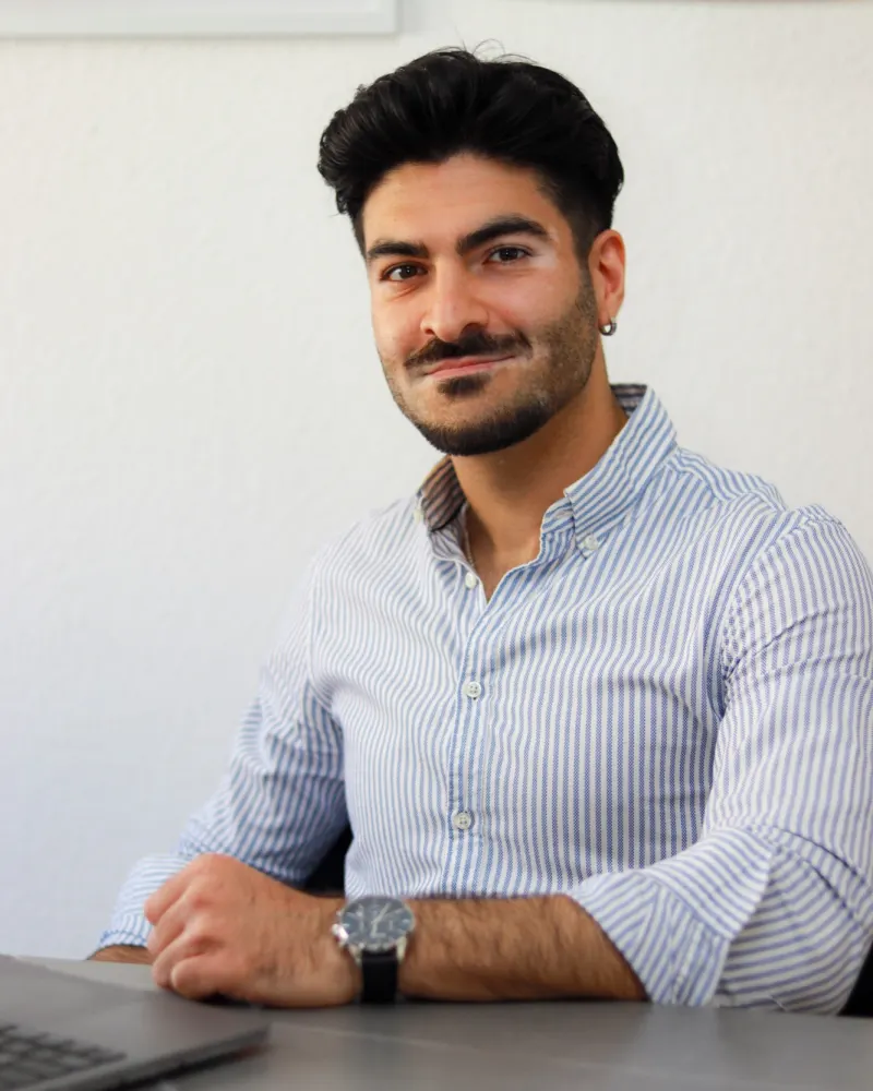 Karuan Osi im blau-weiß gestreiften Hemd, an einem Tisch mit Laptop sitzend, vor heller Wand